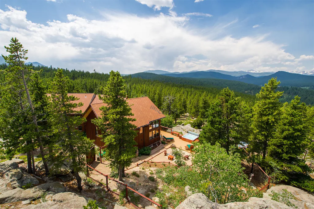 A cabin with its deck and hot tub in the middle of a forest facing mountains