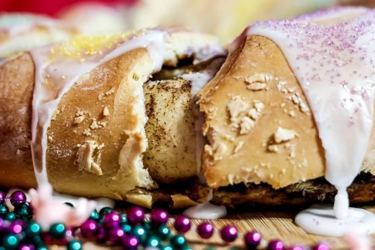 A close up of a glazed King Cake on a wooden board surrounded by colorful Mardi Gras beads